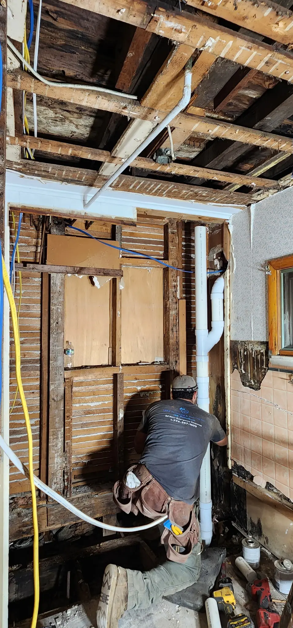 A bathroom stripped back to bare lath and studs, with new white PVC waste pipe and yellow and blue flexible supply lines run through the opened walls, and a technician in a branded 1896 Plumbing & Heating shirt kneeling at the work.