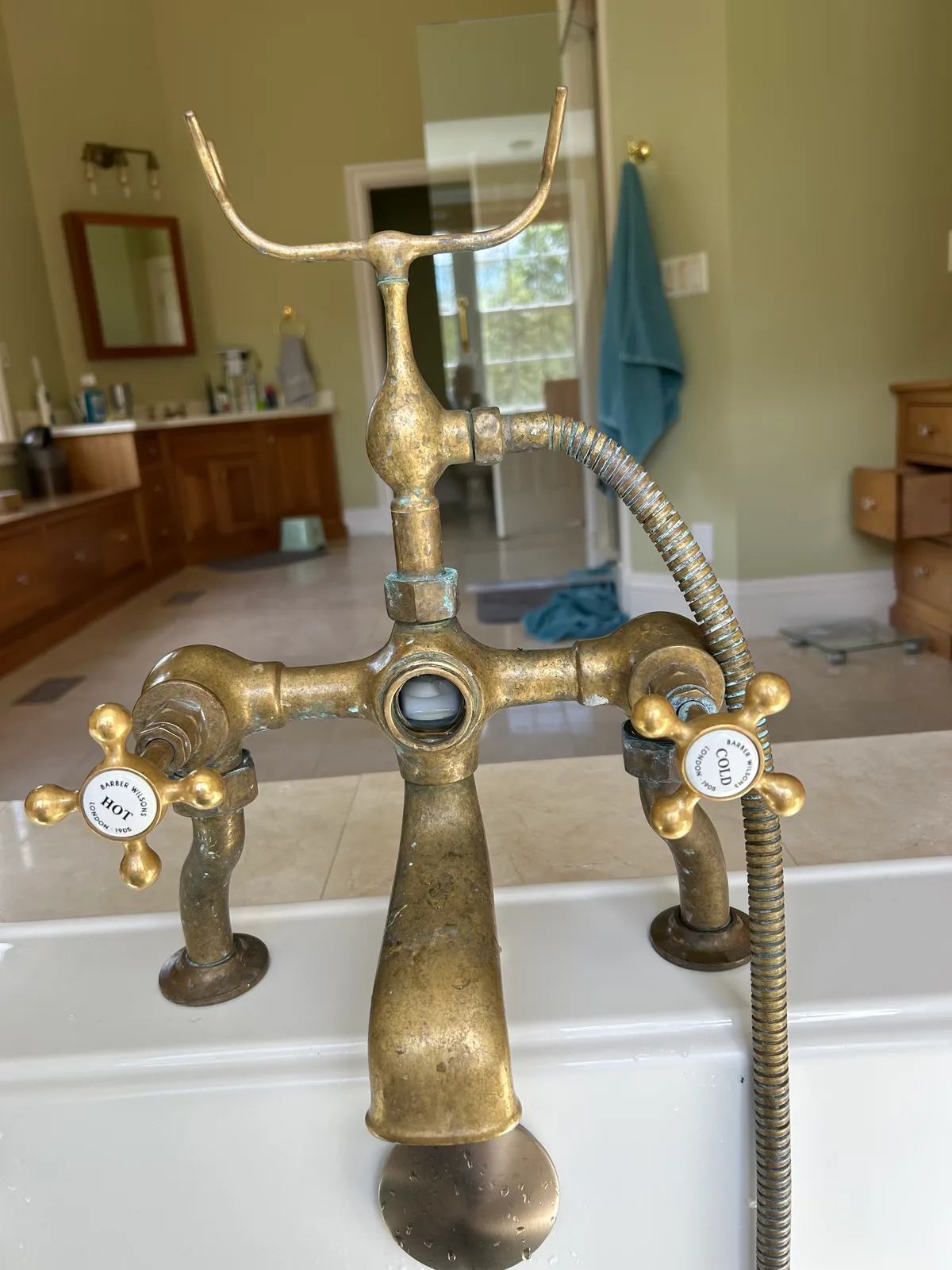 An aged brass tub filler with porcelain-buttoned cross handles marked hot and cold, and a handheld shower on a flexible hose, mounted on the rim of a white bath in a tiled bathroom.