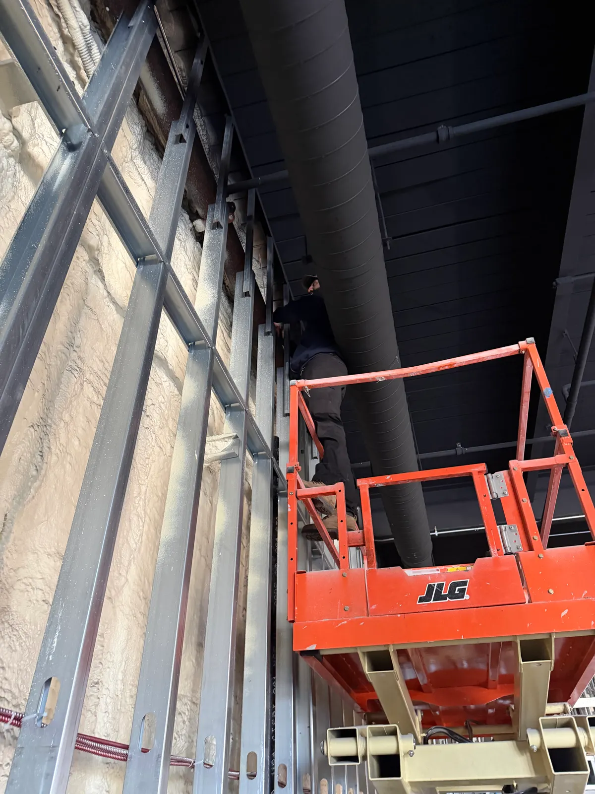 The inside of a large building under construction. Tall steel stud framing runs up a rough stone foundation wall that has been spray-foamed, and a black-painted structural deck crosses the top of the frame with a large spiral-seam duct running through it. A worker stands on the platform of an orange scissor lift, working at the top of the studs.