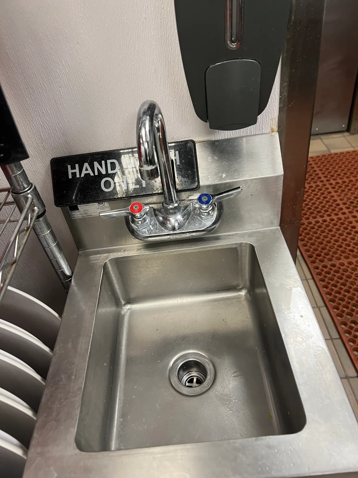 A stainless steel hand-wash sink with a chrome gooseneck faucet and a wall-mounted soap dispenser in a restaurant kitchen, with red anti-fatigue matting and quarry tile flooring alongside it.