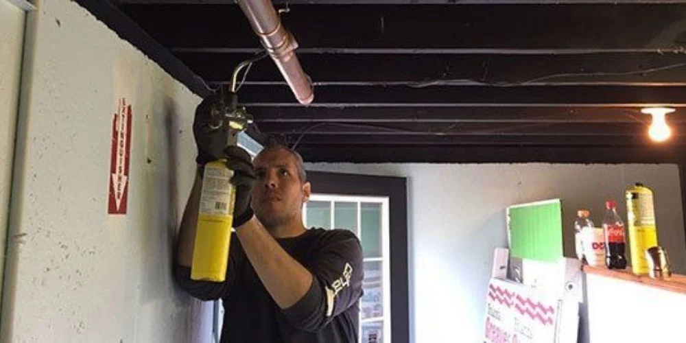 A plumber in a branded work shirt reaching up to fit a valve on overhead copper piping in a commercial basement, beside an exposed joist ceiling.