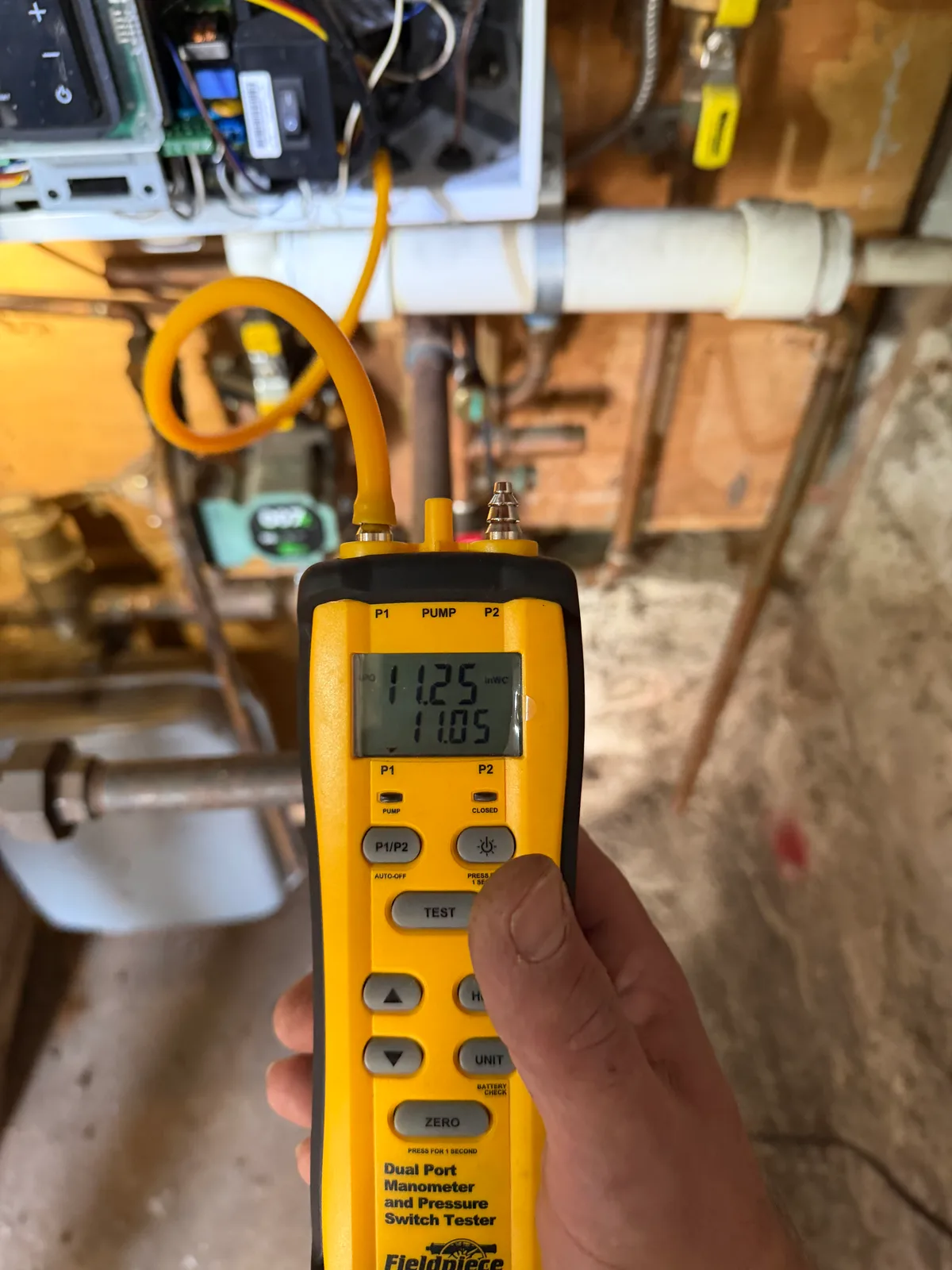 A hand holding a yellow Fieldpiece dual-port manometer in front of an opened appliance control panel, the instrument screen reading 11.25 and 11.05 inches of water column.