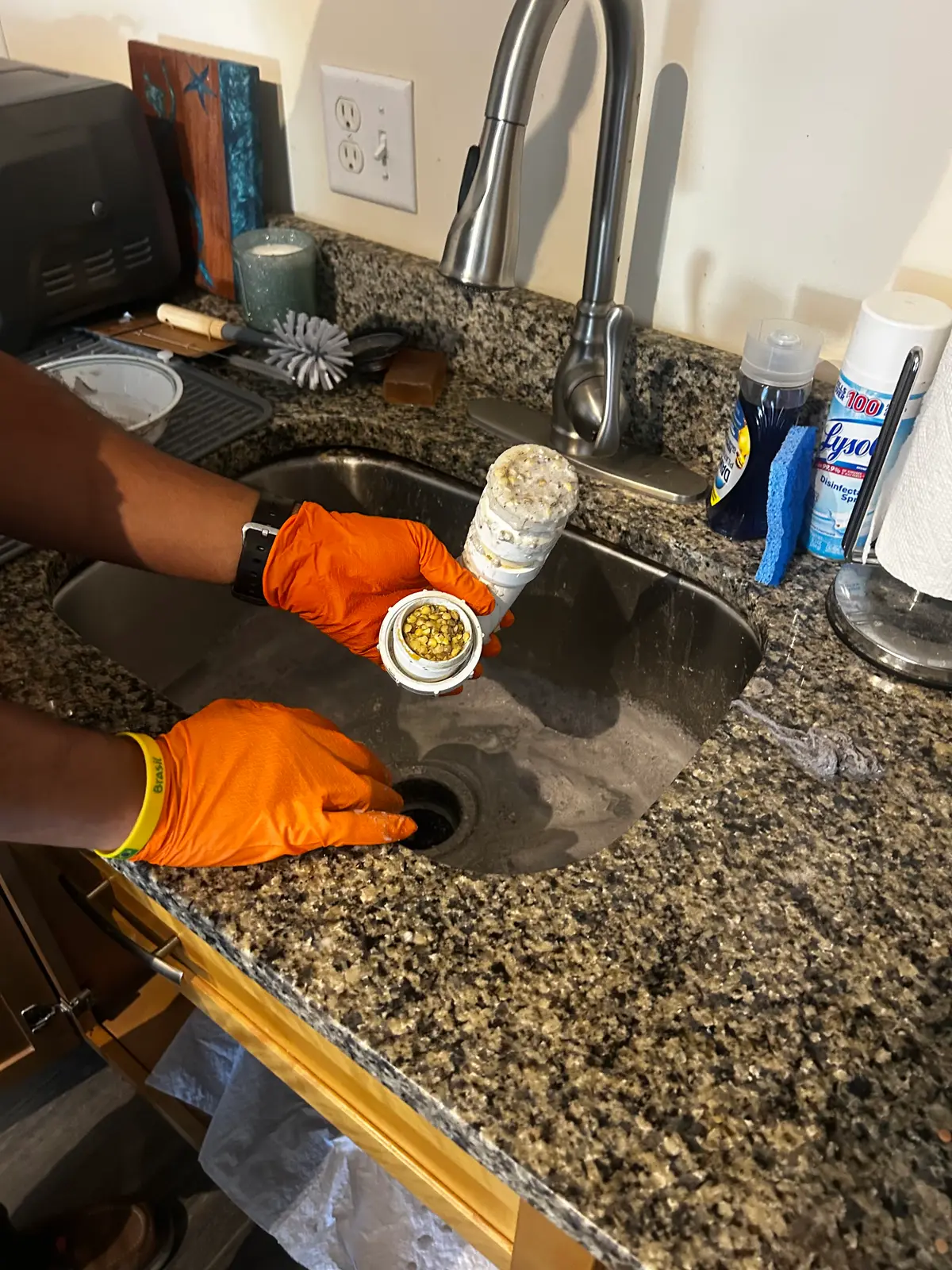 Gloved hands holding a short length of removed kitchen drain pipe packed solid with congealed grease and food waste, over a stainless steel sink set into a granite worktop.