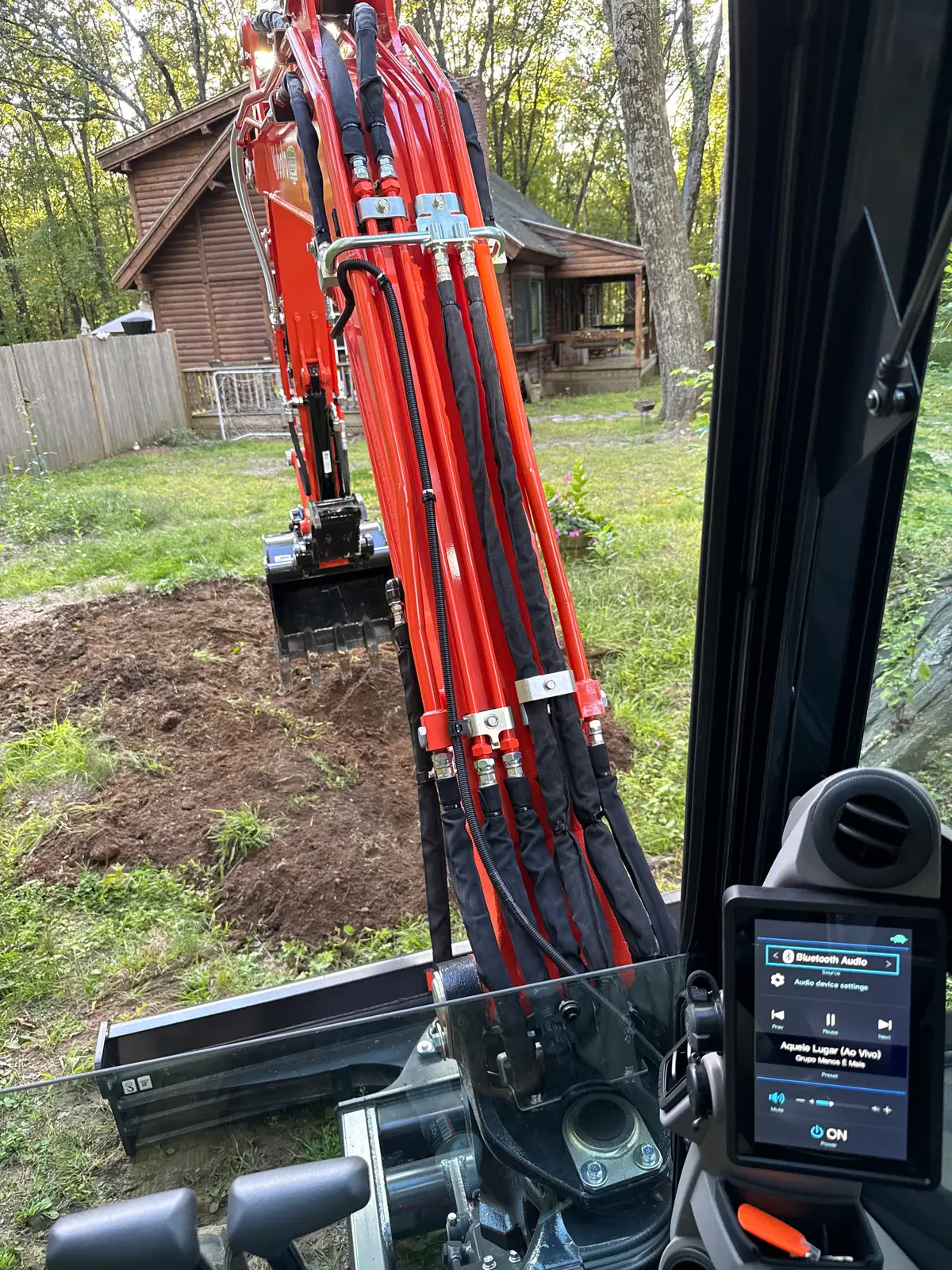 The view from inside the cab of a mini-excavator, out over the machine’s orange boom at a freshly dug trench running across the lawn of a house in woodland.