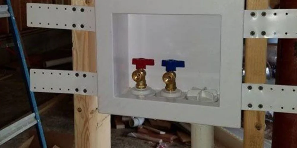 A washing machine outlet box roughed into an open stud wall, with red and blue quarter-turn valves fitted and the waste connection below.