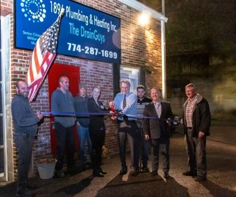 Eight people standing outside the 1896 Plumbing & Heating shop at night for a ribbon cutting, in front of the illuminated company sign, with an American flag on the wall beside them.