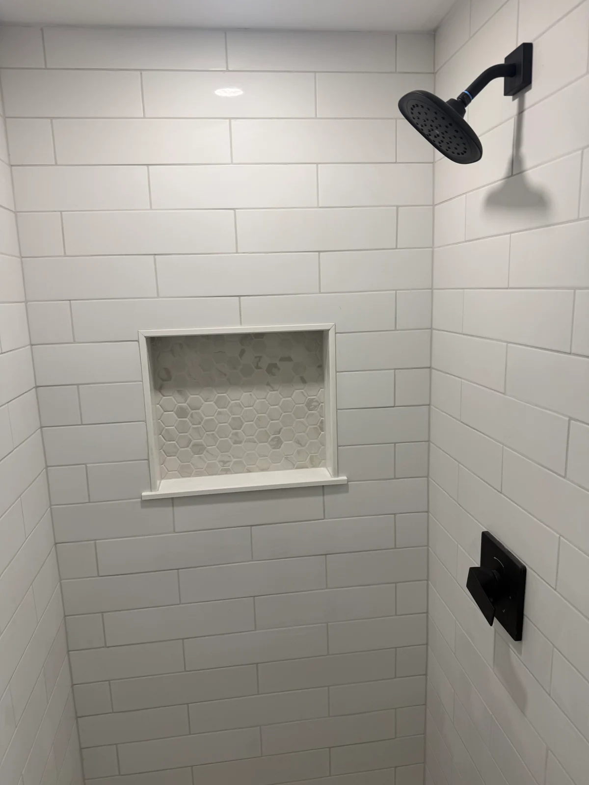 A finished tiled shower. A matte black shower head on a squared arm and wall flange sits high on the right-hand wall, with the matching matte black mixer trim and lever handle below it. A recessed niche with a marble hexagon mosaic back is set into the tiled wall opposite.