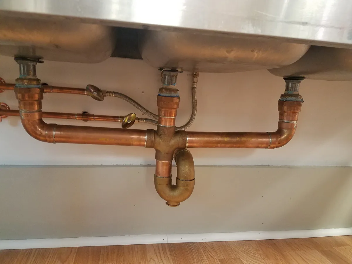 The underside of a three-compartment stainless steel sink, showing new copper waste arms dropping from each bowl into a horizontal run that turns down into a cast brass P-trap, with braided supply connectors and stop valves above.