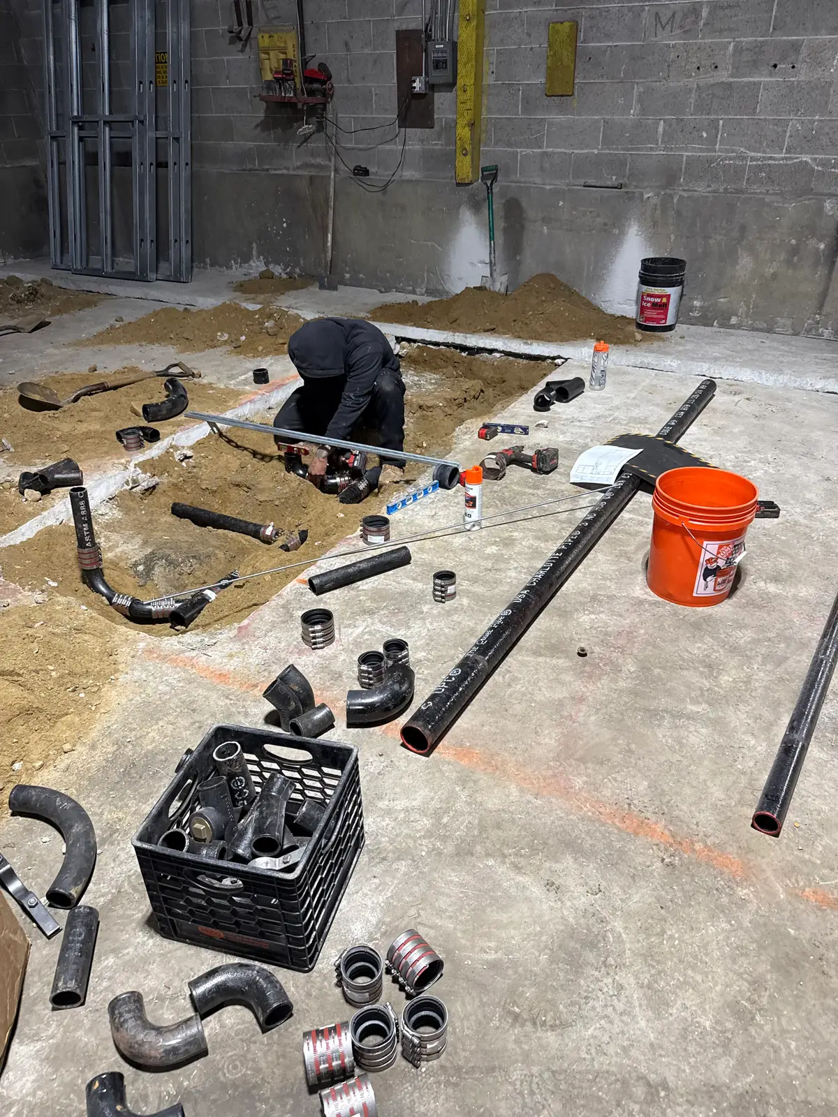 A worker kneeling in an opened cut in a concrete slab, assembling cast-iron underslab drainage, with lengths of soil pipe and shielded no-hub couplings laid out on the concrete around him and steel stud framing behind.