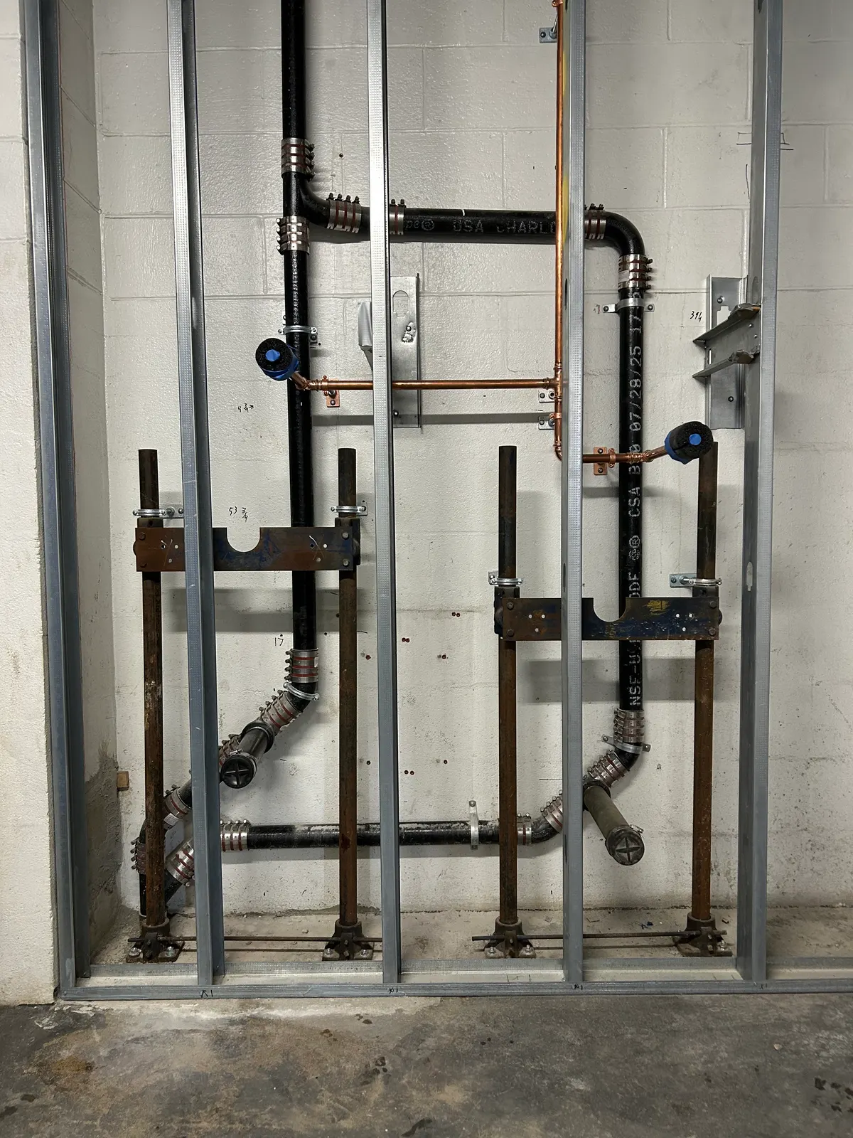 A commercial plumbing rough-in in an unfinished steel-stud wall against a painted concrete block wall. Two matching groups of black cast-iron waste pipe run down to the floor, joined by banded stainless no-hub couplings, with copper water lines crossing at waist height and copper stub-outs clipped to the block. Rusted steel fixture carriers with blue-painted cross plates are bolted to the concrete floor for wall-hung fixtures, and black test caps are still fitted to the waste stubs. Pencil measurements are written on the wall.